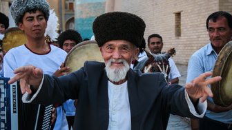 Ein älterer Mann mit einem breiten Lächeln und traditioneller Kleidung tanzt fröhlich in einer Gruppe von Musikern.