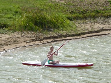 Ein Junge paddelt fröhlich auf einem Stand-Up-Paddle-Board in einem ruhigen Gewässer, umgeben von grüner Natur.