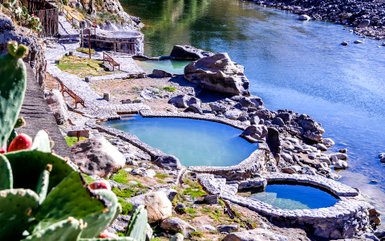 Natürliche Thermalquellen mit klarem Wasser, umgeben von Felsen und einer ruhigen Flusslandschaft im Hintergrund.