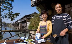 Ein fröhliches Paar steht am Ufer eines malerischen Flusses und präsentiert eine Auswahl an köstlichen Gerichten.
