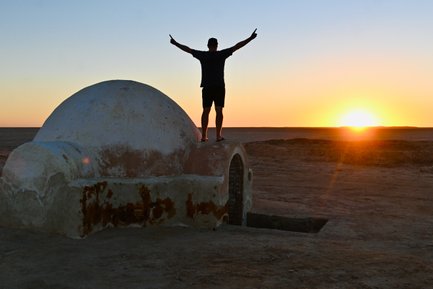 Ein Mann steht auf einem alten, runden Gebäude und breitet seine Arme aus, während die Sonne am Horizont aufgeht.