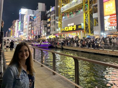 Eine Frau mit Brille lächelt am Ufer des Dotombori-Flusses, umgeben von bunten Neonlichtern und Menschenmengen.