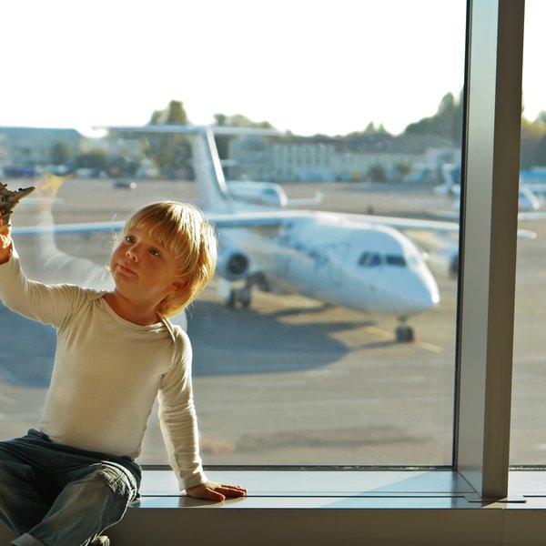 Ein kleiner Junge sitzt am Fenster eines Flughafens und hält ein Spielzeugflugzeug in der Hand, während er nach draußen schaut.