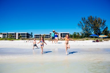 Drei Kinder laufen fröhlich am Strand entlang, während ihre Familie im Hintergrund entspannt. Strahlend blauer Himmel über ihnen.