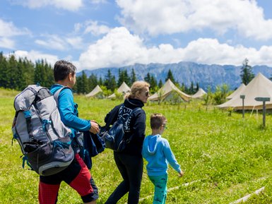 Eine Familie wandert durch eine grüne Wiese, umgeben von Zelten und majestätischen Bergen unter einem blauen Himmel.