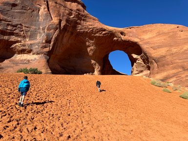 Zwei Personen wandern über eine sandige Landschaft in Monument Valley, umgeben von beeindruckenden roten Felsen und blauem Himmel.