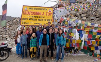 Eine Gruppe von Reisenden posiert fröhlich vor dem Khardungla-Pass-Schild, umgeben von bunten Gebetsfahnen.