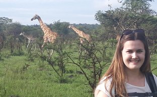 Eine Frau mit Rucksack lächelt vor einer Gruppe von Giraffen, die in der grünen Savanne von Mburo Nationalpark grasen.
