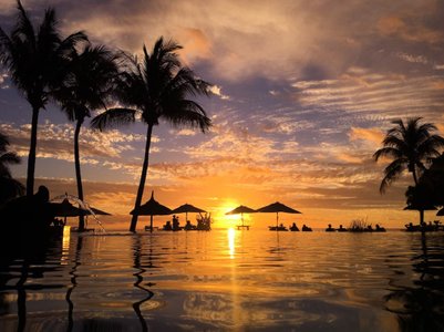 Ein atemberaubender Sonnenuntergang über einem Pool, umgeben von Palmen und Silhouetten von Menschen unter Sonnenschirmen.