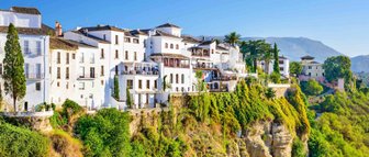 Die weißen Gebäude von Ronda thronen majestätisch auf einer Klippe, umgeben von üppigem Grün und strahlend blauem Himmel.
