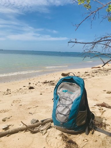 Ein blauer Rucksack steht einsam am Strand, umgeben von feinem Sand und dem glitzernden Wasser des Ozeans.