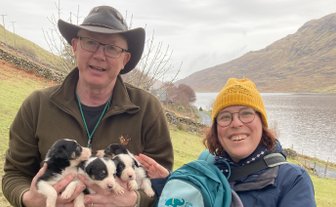 Ein Mann mit einem Hut hält mehrere süße Welpen in seinen Armen, während eine Frau mit einer gelben Mütze lächelt.
