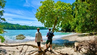 Zwei Wanderer erkunden die Küstenlinie eines tropischen Strandes, umgeben von dichter Vegetation und klarem Wasser.