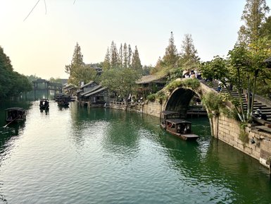Eine ruhige Wasserstraße in Wuzhen, umgeben von traditionellen Holzhäusern und Bäumen, mit Booten, die sanft auf dem Wasser treiben.