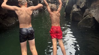 Zwei Jungen springen fröhlich in ein klares, grünes Wasser zwischen hohen, steilen Felsen.