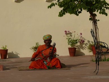 Ein indischer Straßenmusiker sitzt im Freien, trägt ein leuchtendes orangefarbenes Gewand und spielt ein traditionelles Instrument.