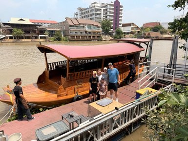 Eine Gruppe von fünf Personen posiert lächelnd vor einem traditionellen Longtailboot am Chao Phraya Fluss.