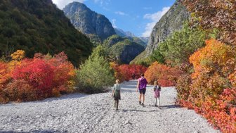 Drei Personen wandern auf einem Kiesweg durch eine farbenfrohe Herbstlandschaft mit Bergen im Hintergrund.