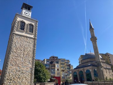 Eine steinerne Uhrturm mit einem Ziffernblatt und ein Minarett stehen unter einem klaren blauen Himmel in Korça.