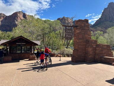 Besucher stehen an der Eingangsstation des Zion Nationalparks, umgeben von beeindruckenden roten Felsen und grünem Laub.