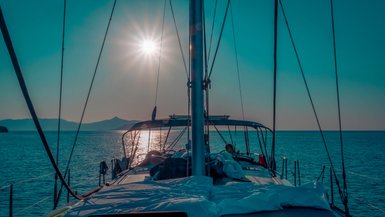 Ein Segelboot fährt über ruhiges Wasser, während die Sonne hell am Himmel strahlt und die Landschaft im Hintergrund sichtbar ist.
