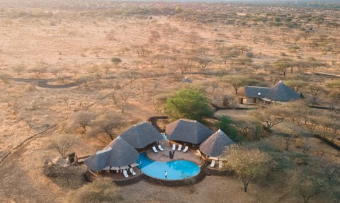 Eine Luftaufnahme eines luxuriösen Safari-Camps mit einem Pool, umgeben von trockener Savanne und sanften Hügeln im Hintergrund.