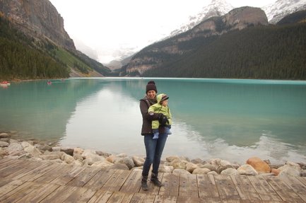 Eine Frau steht am Ufer eines klaren Sees in Kanada und hält ein Baby in einem grünen Anzug in ihren Armen.