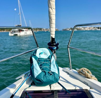 Ein türkisfarbener Rucksack liegt auf dem Deck eines Segelboots, während im Hintergrund andere Boote und eine Küstenlinie sichtbar sind.