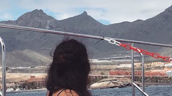 Eine Frau mit langen, dunklen Haaren sitzt auf einem Boot und blickt auf die Berge im Hintergrund.