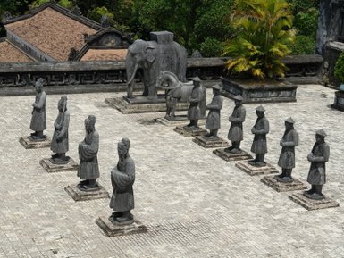 Eine Gruppe von zehn steinernen Figuren steht auf einem Platz, umgeben von üppigem Grün und historischen Gebäuden im Hintergrund.