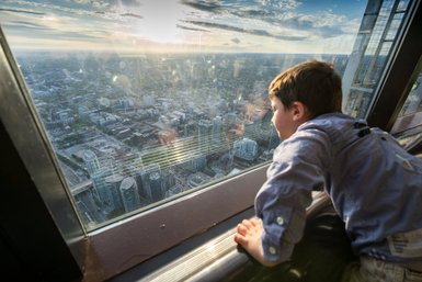Ein Junge schaut fasziniert aus einem hohen Fenster auf die Stadt Toronto, während die Sonne am Horizont untergeht.