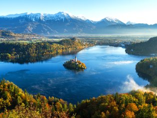 Ein malerischer Blick auf den Bleder See, umgeben von buntem Herbstlaub und majestätischen Bergen im Hintergrund.