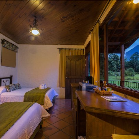 Ein gemütliches Zimmer mit zwei Betten, einem Holztisch und Blick auf die grüne Landschaft und den Vulkan durch große Fenster.