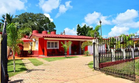 Ein lebhaftes rotes Haus mit einem gepflasterten Weg, umgeben von üppigem Grün und blauen Himmel mit weißen Wolken.