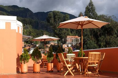 Eine sonnige Terrasse mit Holzmöbeln und Sonnenschirmen, umgeben von grünen Pflanzen und einer beeindruckenden Berglandschaft.