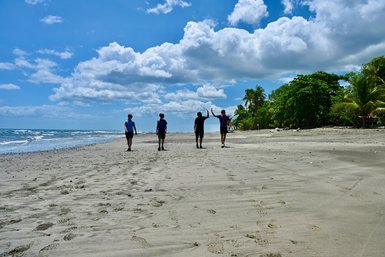 Fünf Menschen gehen entspannt am Strand entlang, während das Wasser sanft gegen das Ufer schlägt und Wolken den blauen Himmel schmücken.