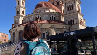 Eine Person mit einem Rucksack steht vor einer beeindruckenden Kirche mit mehreren Kuppeln und einem klaren blauen Himmel.