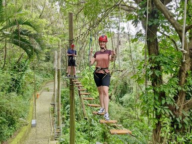 Eine Frau balanciert auf einer Plattform im Kletterpark, umgeben von üppigem Grün und Bäumen.