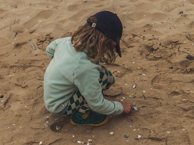 Ein Kind mit lockigem Haar sitzt im Sand und untersucht Muscheln, während es in einem grünen Pullover und gemusterten Hosen gekleidet ist.