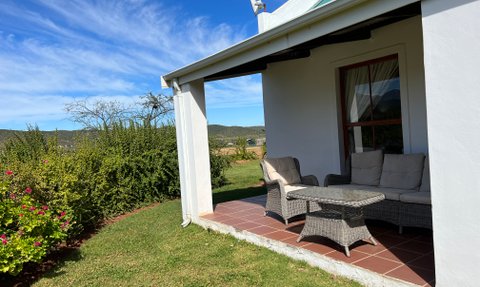 Eine gemütliche Terrasse mit einem Rattansofa und einem Tisch, umgeben von üppigem Grün und blühenden Pflanzen.