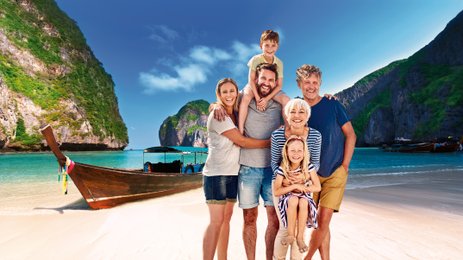 Eine fröhliche Familie posiert am Strand, umgeben von beeindruckenden Klippen und einem Boot im Hintergrund.