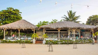 Ein Strandrestaurant mit Strohdach und Palmen, umgeben von Sand, bietet eine entspannte Atmosphäre für Gäste.