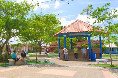 Ein lebhafter Park mit Bänken, wo Menschen entspannen und sich unterhalten, umgeben von üppigem Grün und bunten Gebäuden.