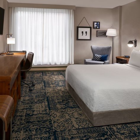Ein modernes Hotelzimmer mit einem großen Bett, einem Schreibtisch und einem gemütlichen Sessel neben einem Fenster mit Vorhängen.