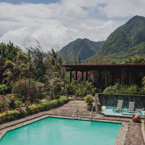 Ein einladender Pool umgeben von üppigem Grün und bunten Blumen, mit Bergen im Hintergrund und einem klaren Himmel.