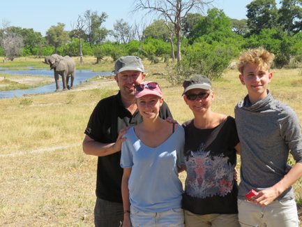 Eine Familie posiert fröhlich vor einem Fluss, während ein Elefant im Hintergrund entspannt grast.