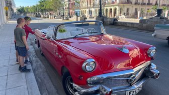 Zwei Jungen stehen neben einem roten Oldtimer-Cabrio, das als Taxi gekennzeichnet ist, auf einer ruhigen Straße.