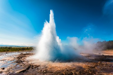 Ein beeindruckender Geysir sprudelt in die Höhe, umgeben von dampfenden Quellen und einer klaren blauen Himmel.