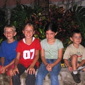 Vier Kinder sitzen fröhlich auf einem großen Stein, umgeben von üppigem Grün und einer gemütlichen Atmosphäre.