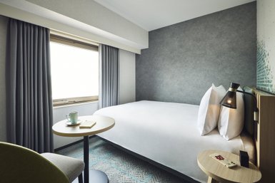 Ein modernes Hotelzimmer mit einem großen Bett, einem kleinen Tisch und einem Fenster, das viel Tageslicht hereinlässt.